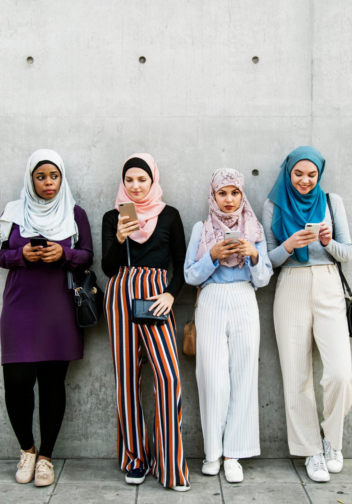 4 women wearing hijabs using smartphones, leaing against a concrete wall