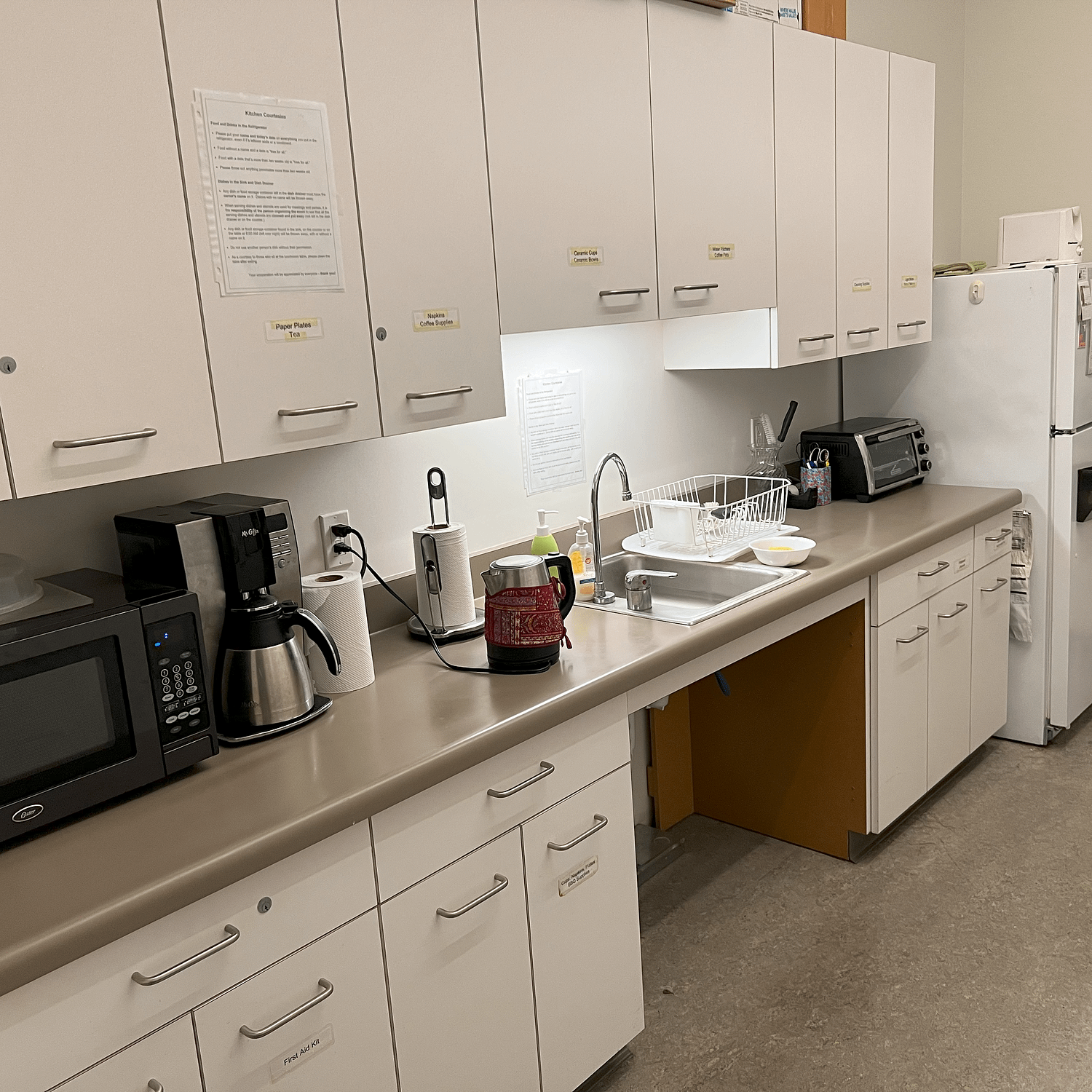 WID's office kitchen, with cabinets, a microwave, coffee maker and electric tea kettle, toaster oven, full size refrigerator, and a kitchen sink with open space underneath the sink to allow a wheelchair user to use the sink comfortably