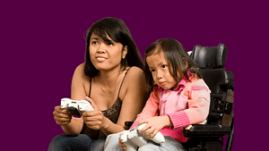 An Asian mother sitting on a couch and daughter sitting in a power wheelchair playing video games.