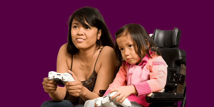 An Asian mother sitting on a couch and daughter sitting in a power wheelchair playing video games.