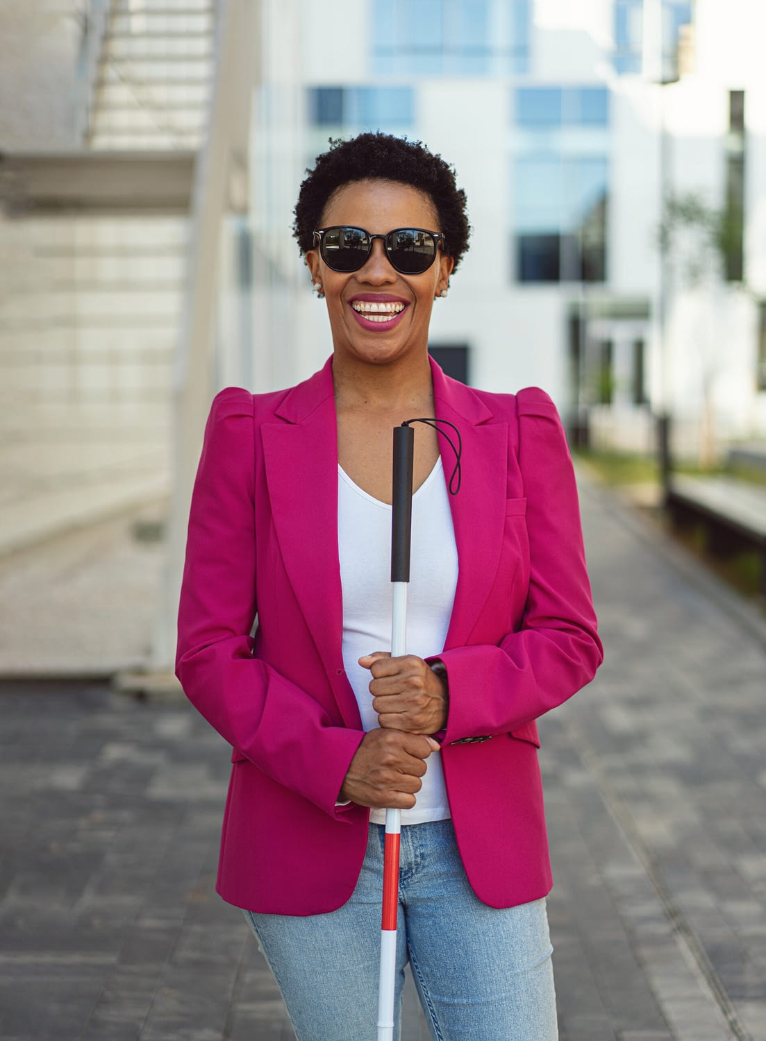 Decorative: A mixed race woman wearing sunglasses and using a white cane. She is smiling and wearing a pink blazer.