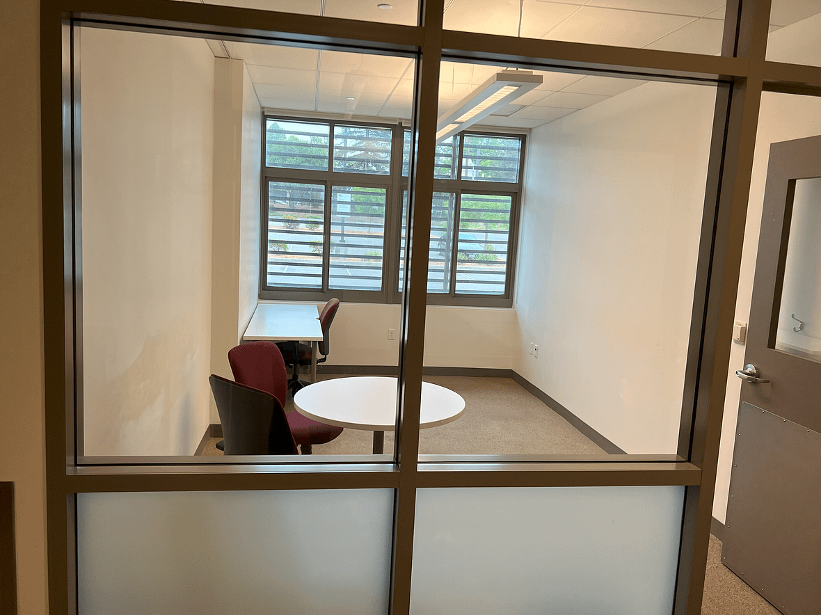 Large office with windows looking out on the parking lot. The room easily fits multiple desks and a small table with two chairs. Two large interior windows bring the light from the outside into the WID office hallway.