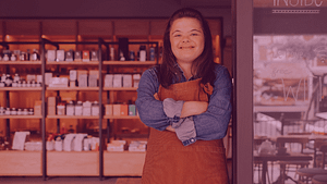 A young woman with Down Syndrome smiles while wearing a long-sleeved shirt and apron with her arms crossed in a coffee shop.