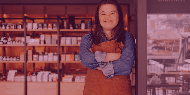 A young woman with Down Syndrome smiles while wearing a long-sleeved shirt and apron with her arms crossed in a coffee shop.