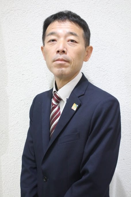 Naoki Kurano, a Japanese man wearing a dark blue suit and red striped tie, stands infront of a white wall and is looking at the camera. 