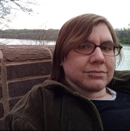 Author photo of Kale Sastre, a white person with short brown hair, glasses, and a lip ring, sitting in front of water and trees.