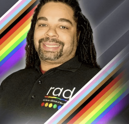 Roy Jones smiles while wearing a black polo shirt with "RAD" printed on it. Rainbows are in the border of this photo.