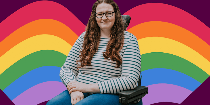 Shona Louise a white woman with long wavy brown hair and a white blouse with blue swipes and jeans sits in her power wheelchair. A Pride rainbow is behind her.