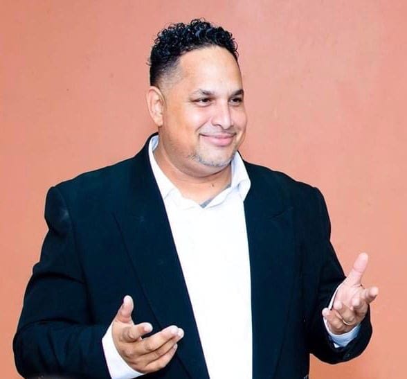 Bryan Rodrigues, a Trinbagonian man with light skin and curly black hair, wearing a navy suit.