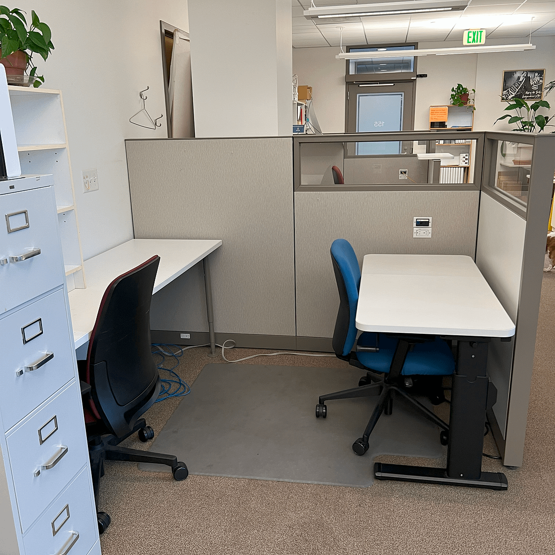 Middle cubicle with two desks and two chairs, a small bookshelf, and a full sized filing cabinet