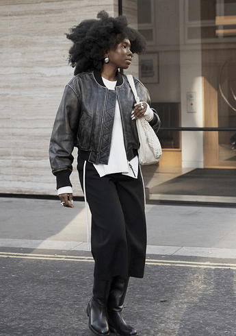 Danielle Oreoluwa Jinadu poses for a portrait photo standing in the middle of a street and looking to her right. She is wearing a leather jacket, blouse, slacks and boots.
