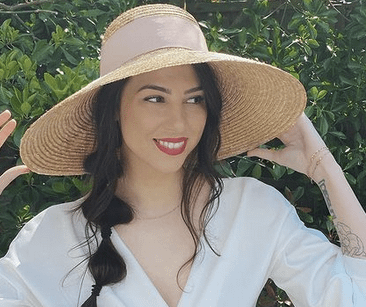 Stevie Boebi smiles looking to her right, wearing a straw sun hat and white blouse.