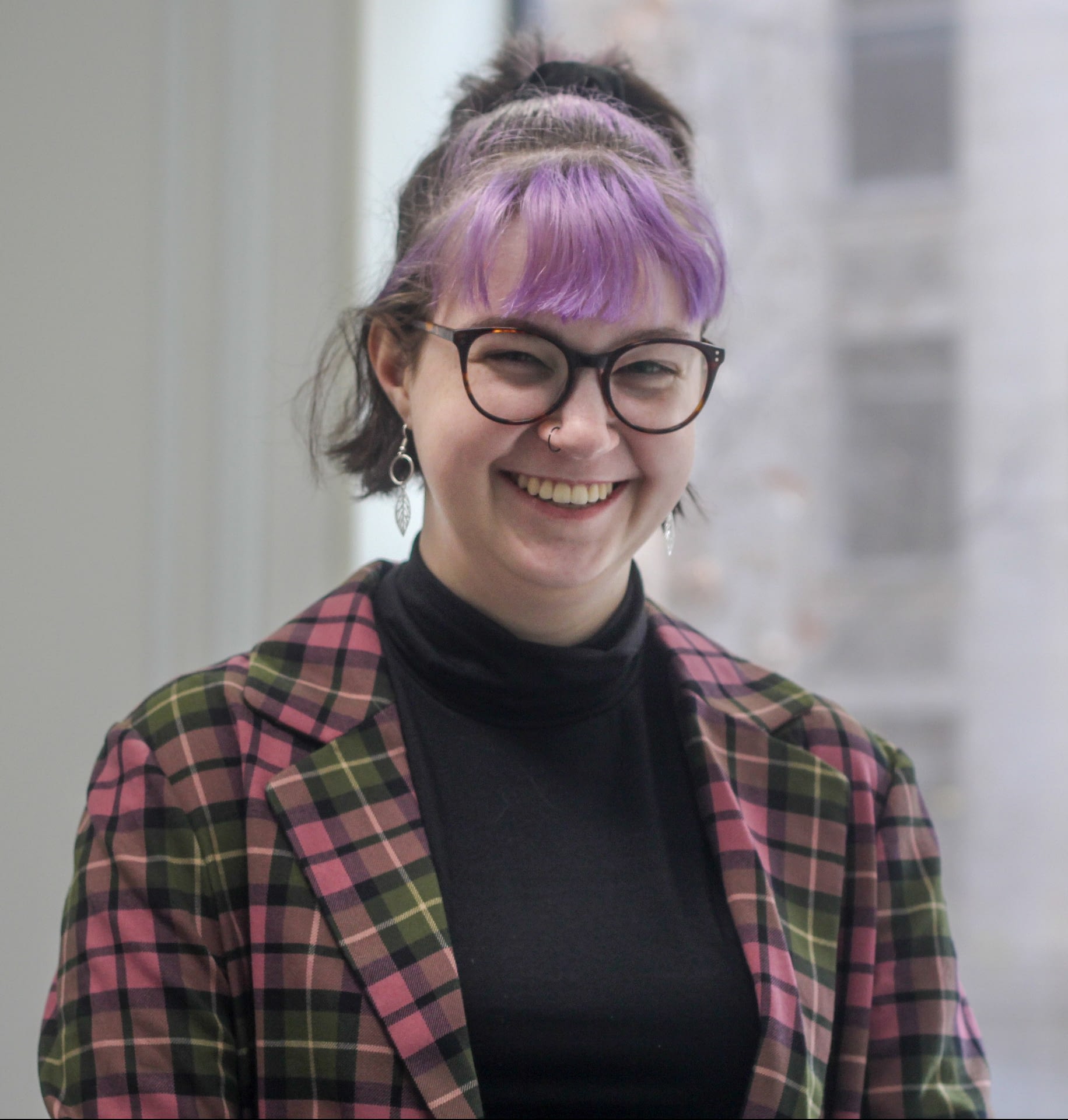 Photo of Issy Orosz, a young white disabled person with short brown and lavender hair. They are wearing a plaid blazer, black turtleneck sweater, and round glasses.