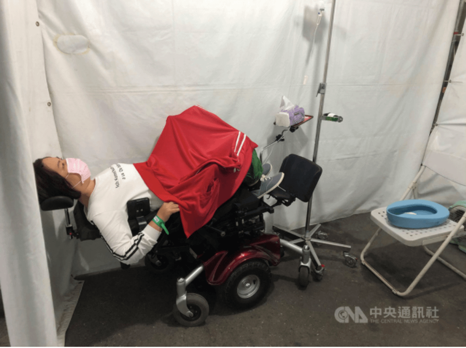 A Taiwanese woman reclining in her electric wheelchair inside a makeshift hospital room inside of a tent. Her jacket is draped over her legs like a blanket.