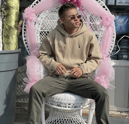 Chella Man looks to his right while wearing sunglasses and a taupe hoodie and pants.