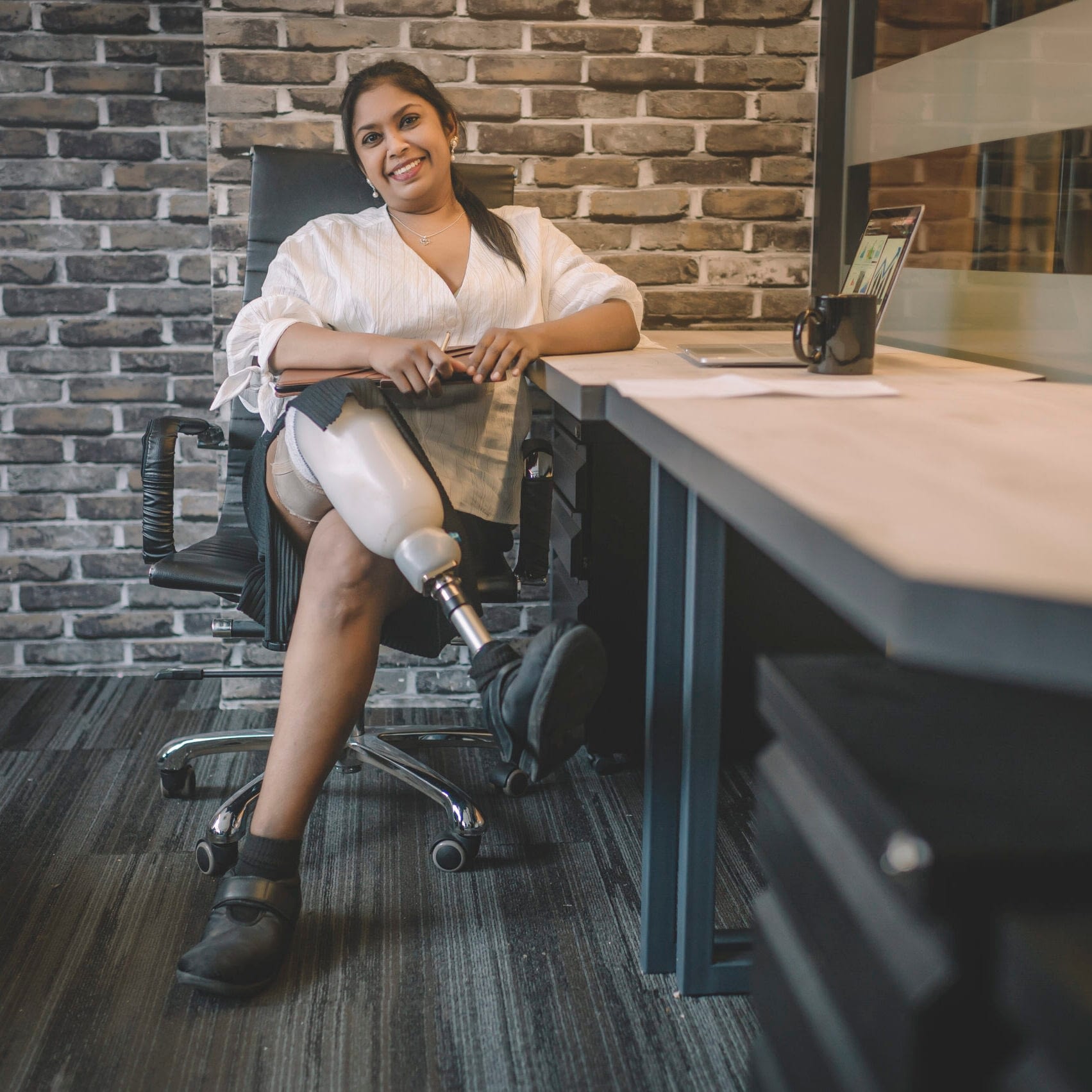 A South Asian woman with brown skin and a prosthetic right leg sits at her desk in an office.