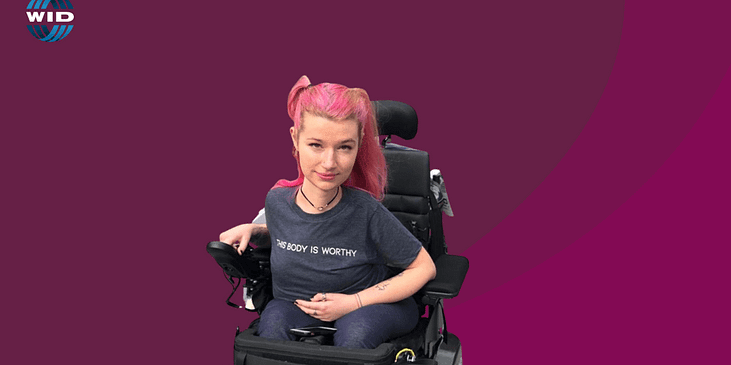 Hannah Soyer, a white woman with pink pig-tailed hair and a gray t-shirt reading “This Body is Worthy” sits in her power wheelchair.