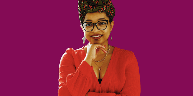 Keri Gray, A Black woman wearing a headwrap, eyeglasses and long sleeved blouse smilles with her left hand on her chin.