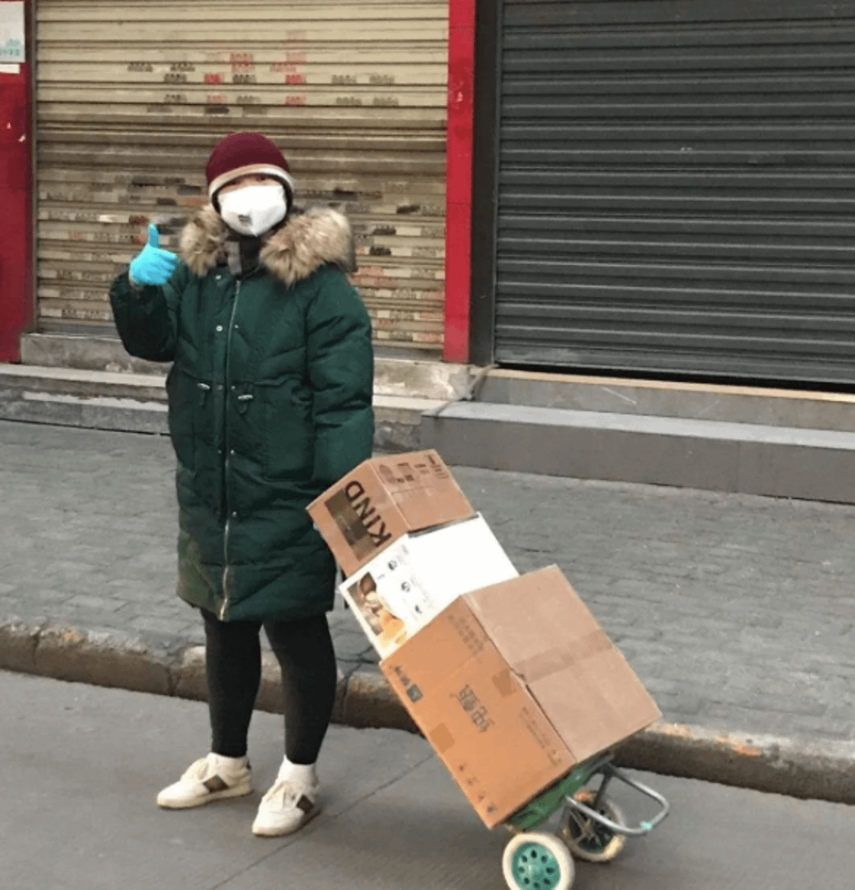 A deaf woman bundled up in a heavy coat, knit hat, disposable gloves, and a face mask pulling boxes of supplies using a small 2-wheeled dolly. She is giving a thumbs up to the camera.