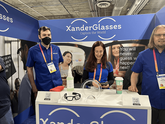 A group of Xanderglasses employees smile at an exhibit with the glasses on a table.