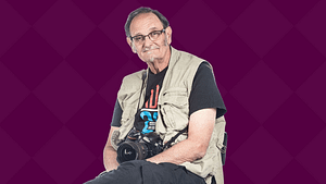 Tom Olin, a White woman with a beige vest and camera around his neck sits on a chair smiling.