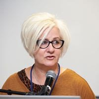 Author photo of Shelley Tremain, a white woman with jaw length white-blonde hair. She is speaking into a microphone.