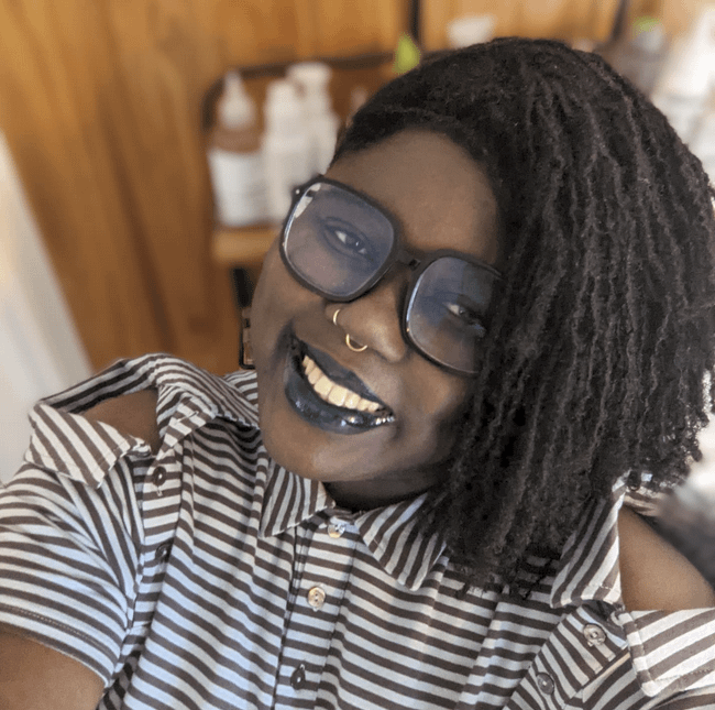 Olu Niyi-Awosusi smiles while wearing black rimmed eyeglasses, a nose ring, black lipstick and a white shirt with black stripes.