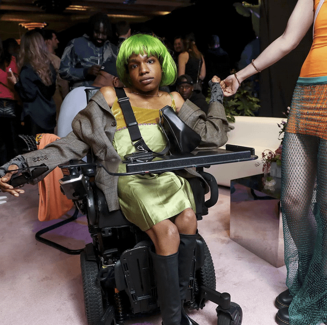 Aaron Rose Phillip poses in her power wheelchair with green hair and a green dress and grayish brown patterned blazer.