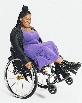Tatiana A Lee smiles while sitting in her wheelchair wearing a leather jacket, a long dress and boots.