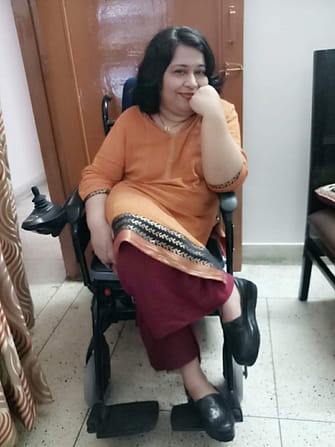 Abha Khetarpal, an Indian woman, sitting in her power chair and smiling. She is wearing a beautiful orange tunic and deep red pants.