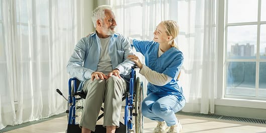 Caregiver in blue scrubs kneeling beside smiling elderly man in wheelchair, sharing warm moment indoors.