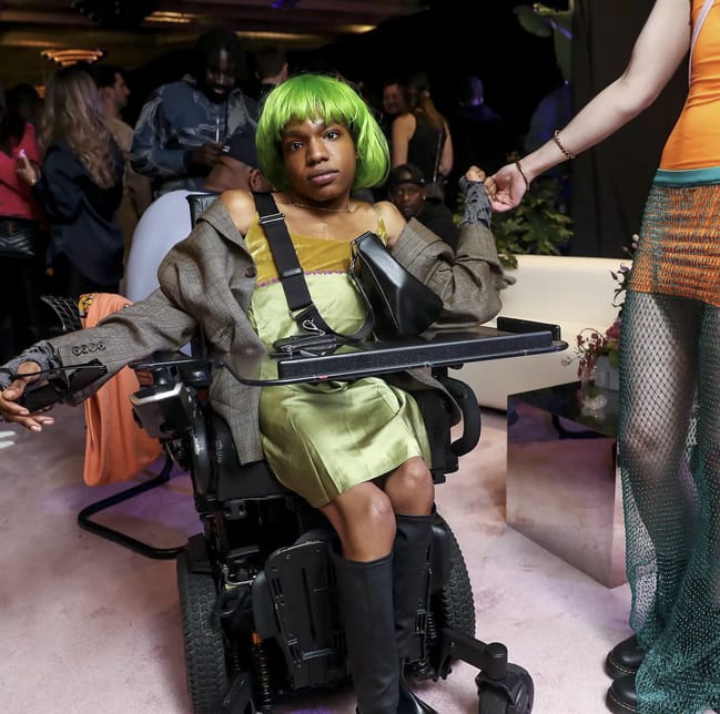 Aaron Rose Phillip poses in her power wheelchair with green hair and a green dress and grayish brown patterned blazer.