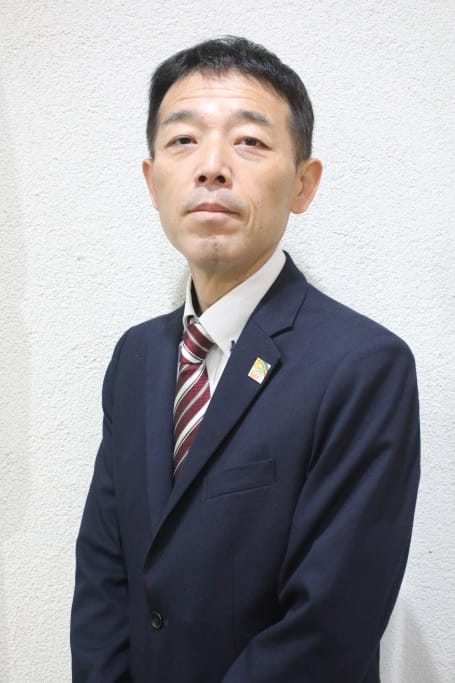 Naoki Kurano, a Japanese man wearing a dark blue suit and red striped tie, stands infront of a white wall and is looking at the camera.