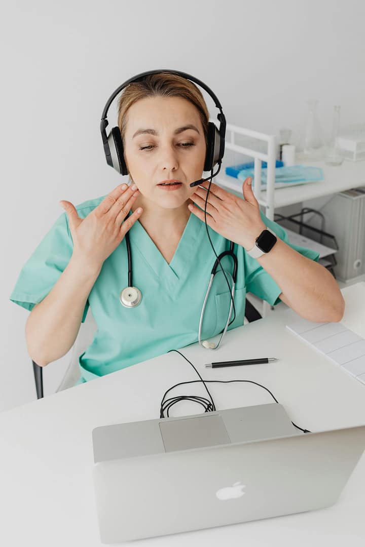 Healthcare provider wearing green scrubs, a headset and communicating using hand gestures in front of a laptop.