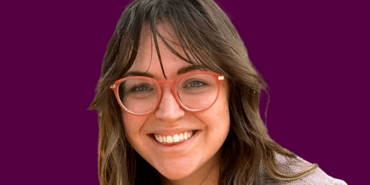 Bri Beck, a White woman of short stature with red-rimmed eyeglasses, a taupe cardigan, black shirt and blue jeans smiles with her right hand on her hip
