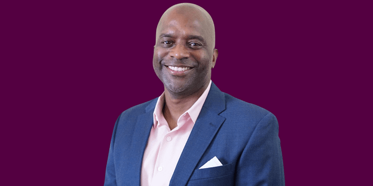 Miguel Quinones, a Black man with a bald head and navy blue suit and pink button down shirt smiles