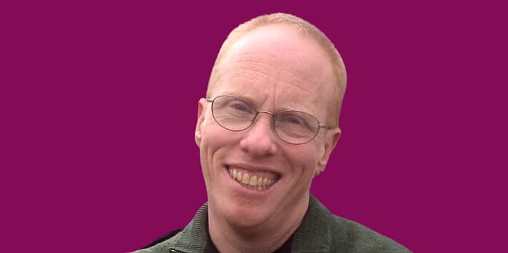 Eli Clare, a white man with eyeglasses and a knit sweater on smiles.