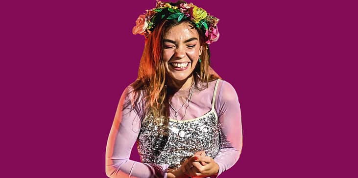 Chloe Hayden smiles with her eyes closed while wearing a flower crown and sparkly dress and long sleeved shirt underneath her dress