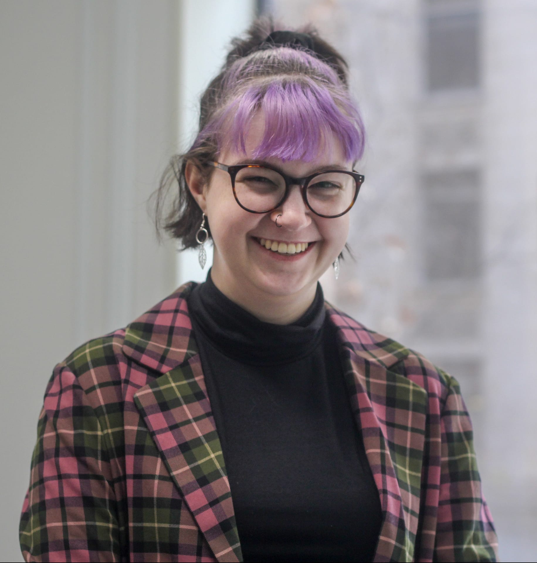 Photo of Issy Orosz, a young white disabled person with short brown and lavender hair. They are wearing a plaid blazer, black turtleneck sweater, and round glasses.