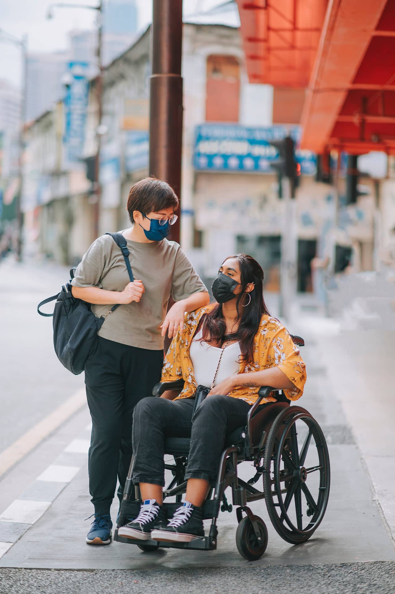 A Chinese woman talking to her friend, an Indian woman with disability using a wheelchair. They are on a sidewalk in a busy city.